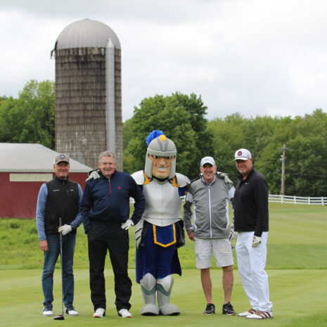 Four men and a person in a knight mascot costume stand on a golf course with a red barn and silo in the background.