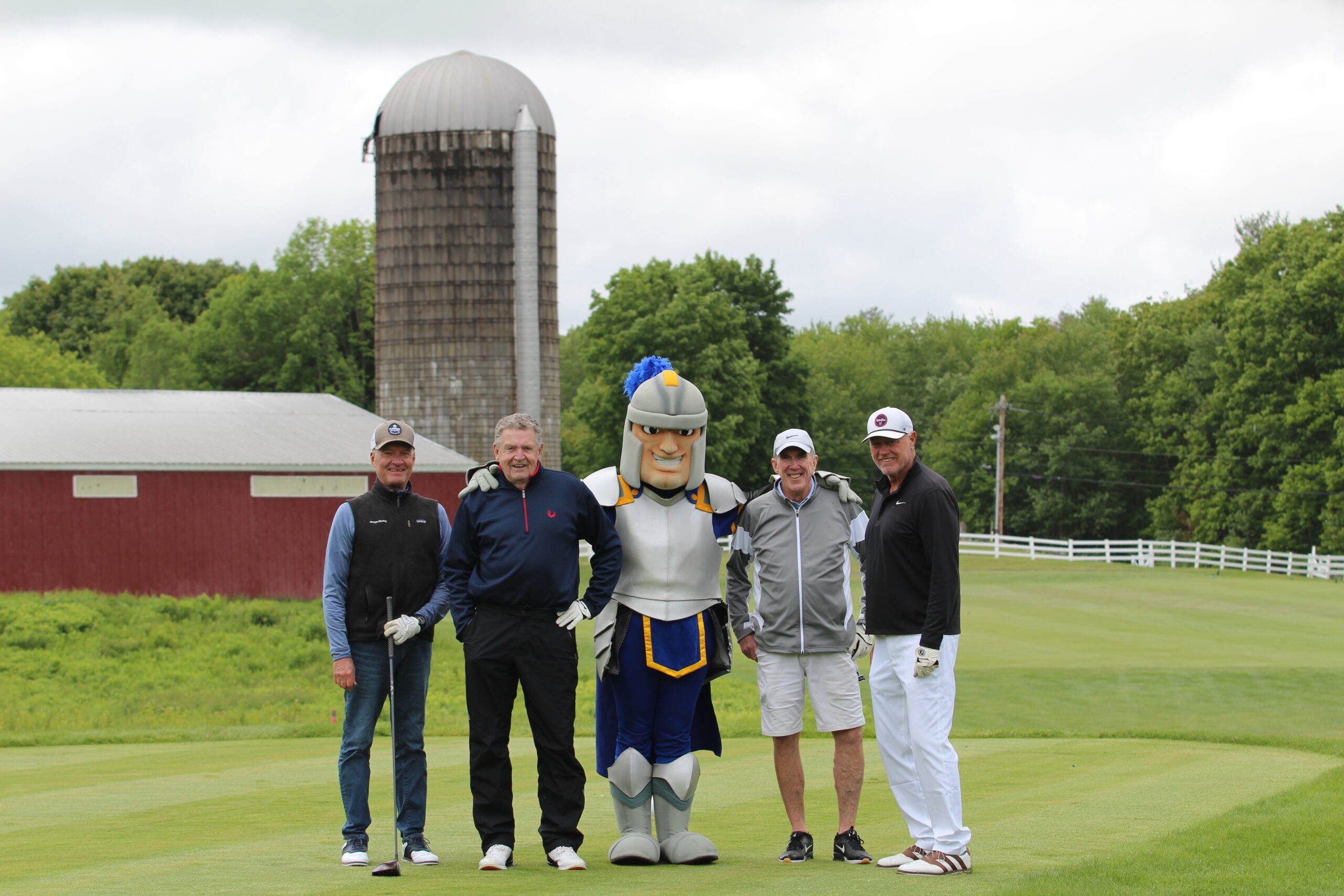 Four men and a person in a knight mascot costume stand on a golf course with a red barn and silo in the background.