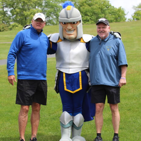 Two men standing on grass with a person in a knight mascot costume, all smiling at the camera on a cloudy day.