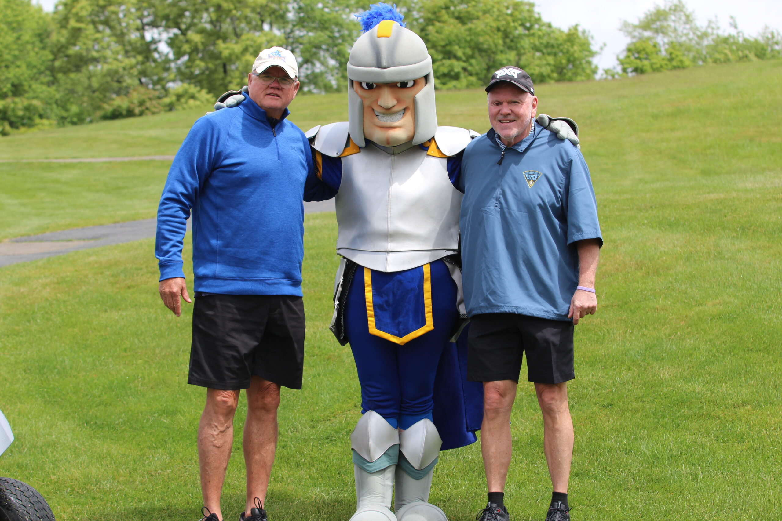 Two men standing on grass with a person in a knight mascot costume, all smiling at the camera on a cloudy day.