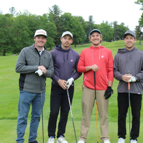 Four men stand on a golf course holding clubs, dressed in casual golf attire with greenery and trees in the background.