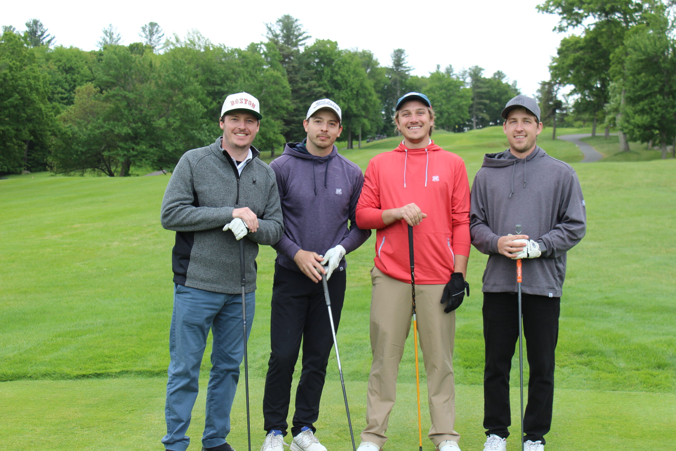 Four men stand on a golf course holding clubs, dressed in casual golf attire with greenery and trees in the background.