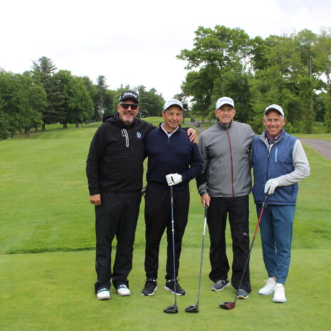 Four men in golf attire stand side by side on a golf course, each holding a golf club, with green grass and trees in the background.