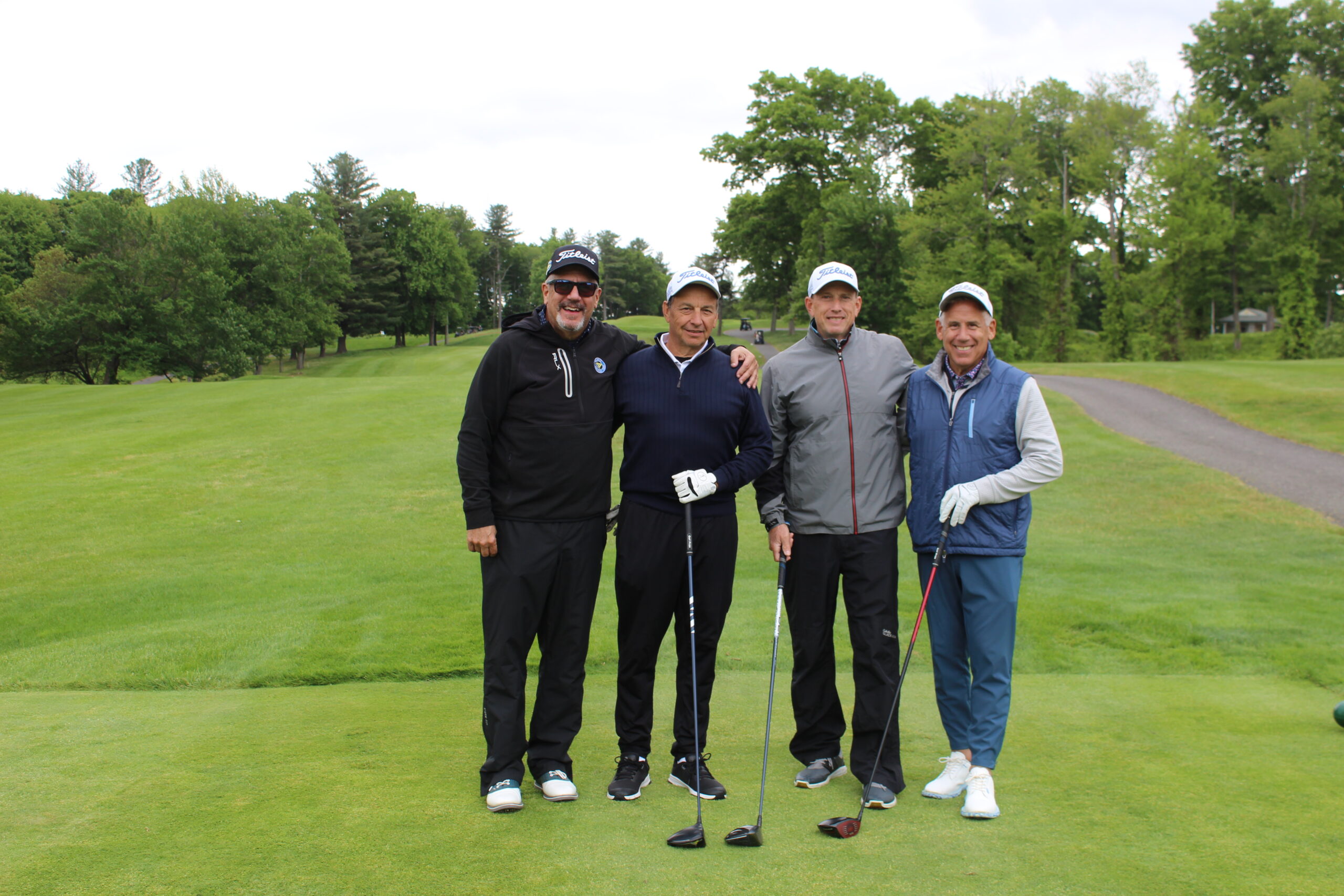 Four men in golf attire stand side by side on a golf course, each holding a golf club, with green grass and trees in the background.