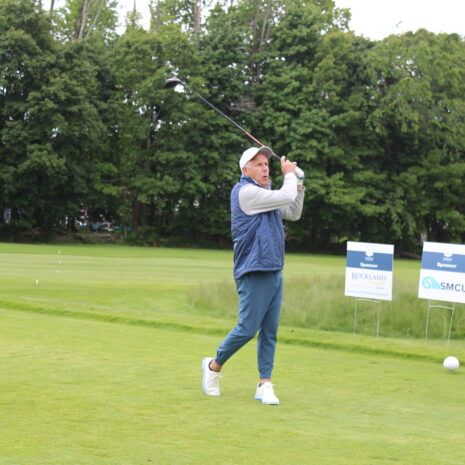 A man in casual golf attire follows through on a golf swing on a green course, with sponsor signs and trees in the background.