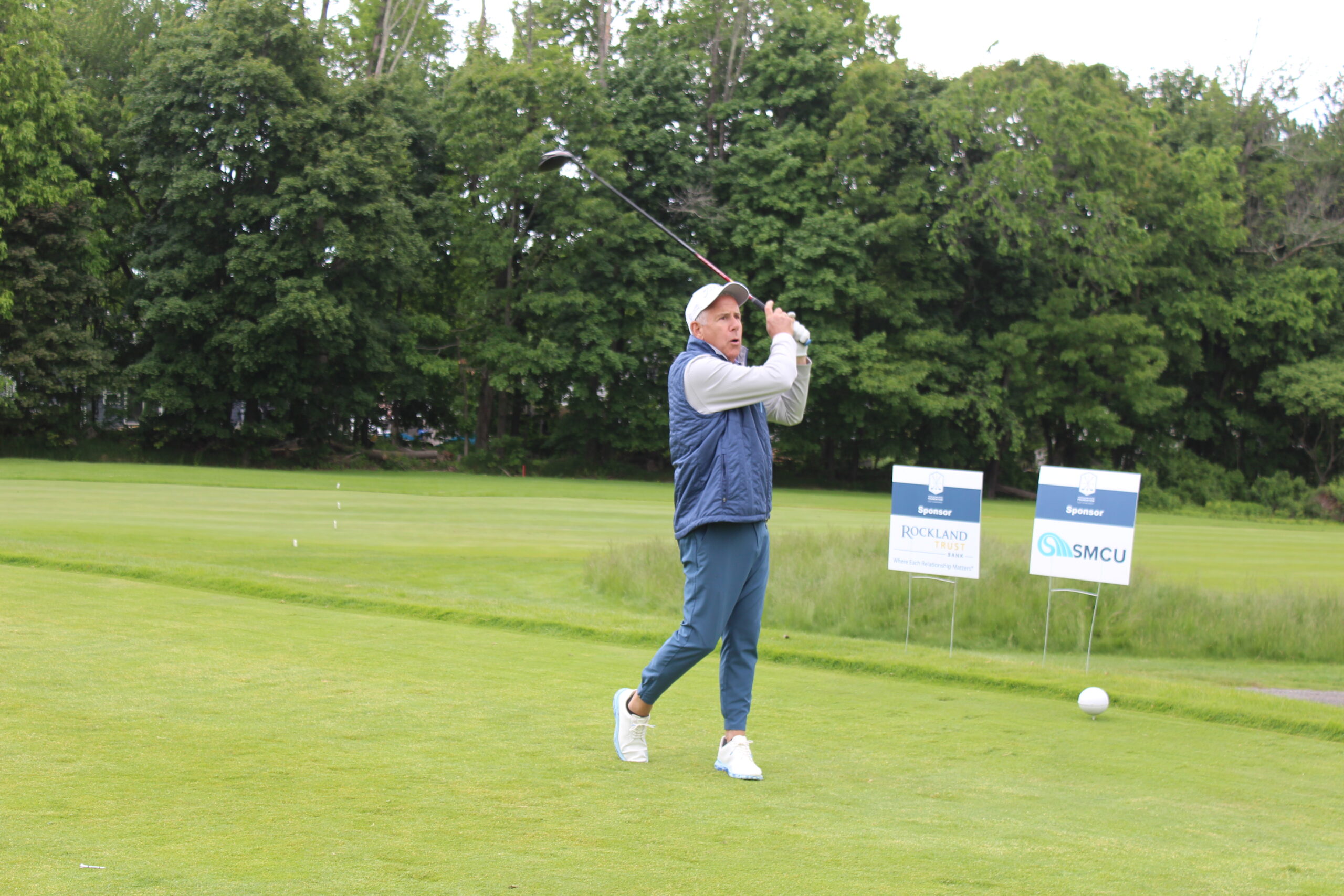 A man in casual golf attire follows through on a golf swing on a green course, with sponsor signs and trees in the background.