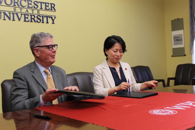 Two people in business attire sit at a table signing documents, with a Worcester State University sign on the wall behind them.