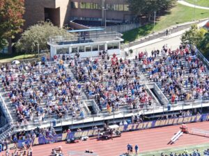 A crowd of people sits and stands in bleachers at an outdoor sports stadium at Fort Hays State University on a sunny day.