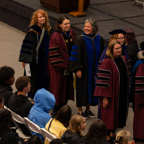A group of faculty in academic regalia stand in a line at a graduation ceremony while students sit in rows of white chairs in the foreground.