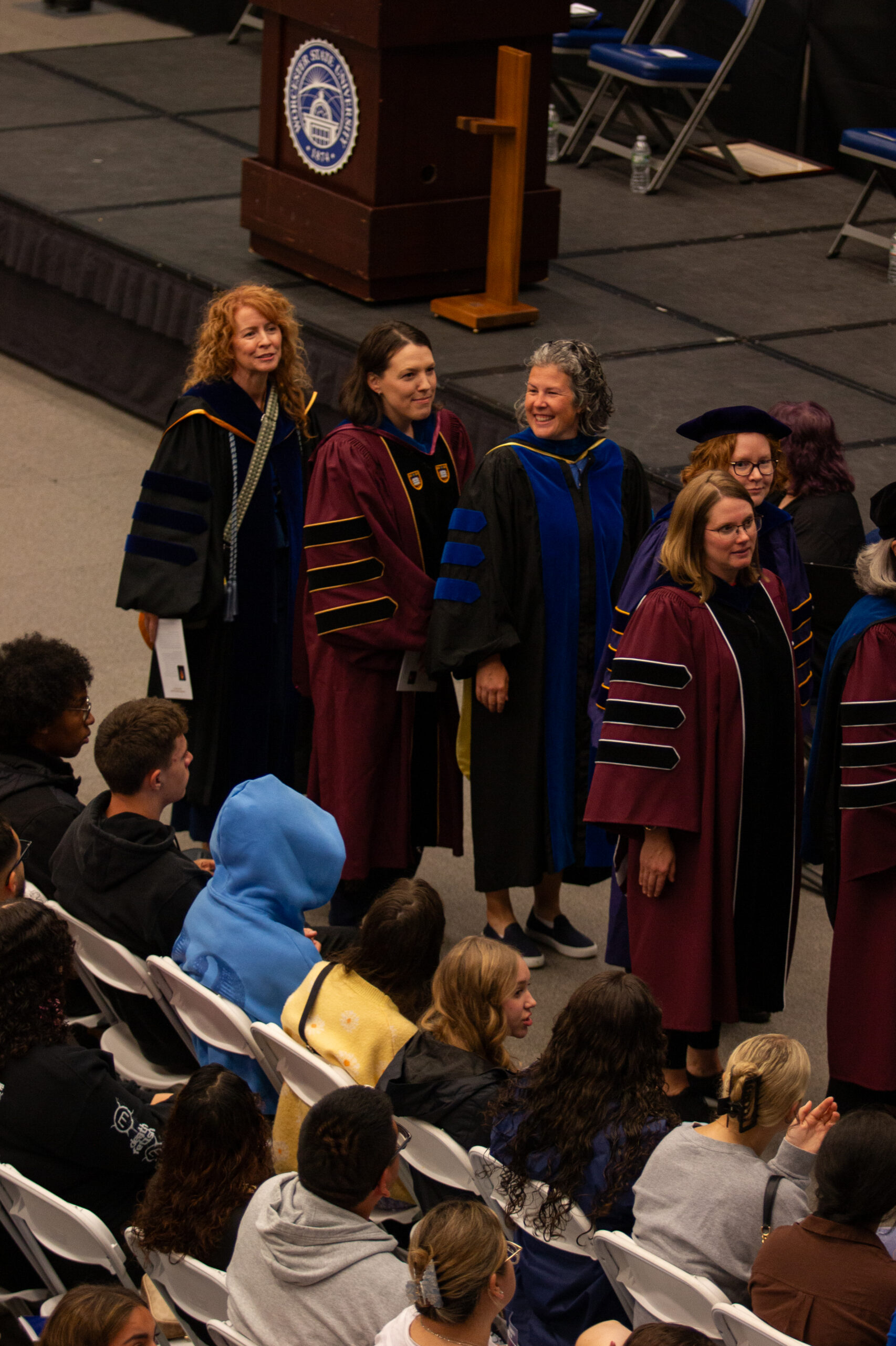 A group of faculty in academic regalia stand in a line at a graduation ceremony while students sit in rows of white chairs in the foreground.