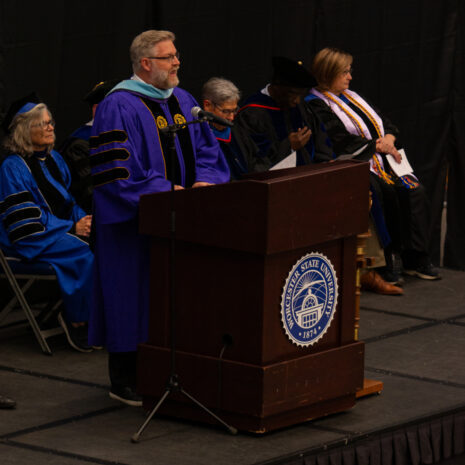 A man in academic regalia speaks at a podium with the Texas A&M University–Commerce seal, while others in robes sit behind him on stage.