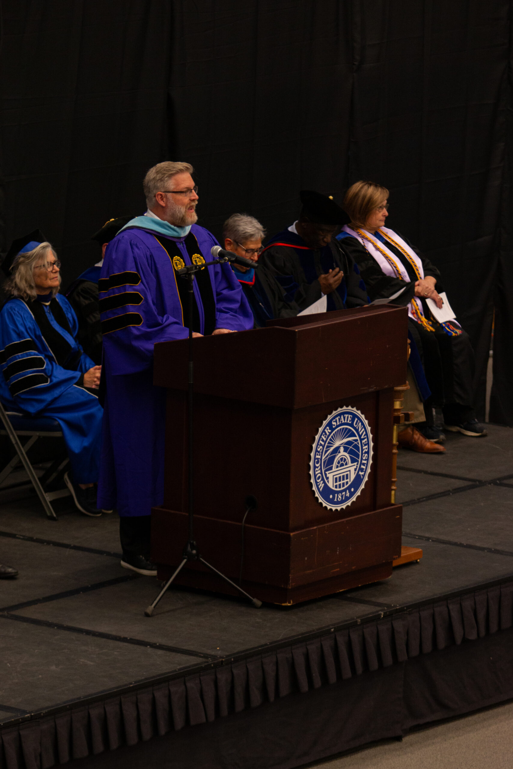 A man in academic regalia speaks at a podium with the Texas A&M University–Commerce seal, while others in robes sit behind him on stage.