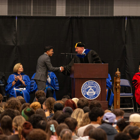 A person in a suit shakes hands with a speaker in academic regalia at a podium during a graduation ceremony, with faculty seated on stage and audience members watching.