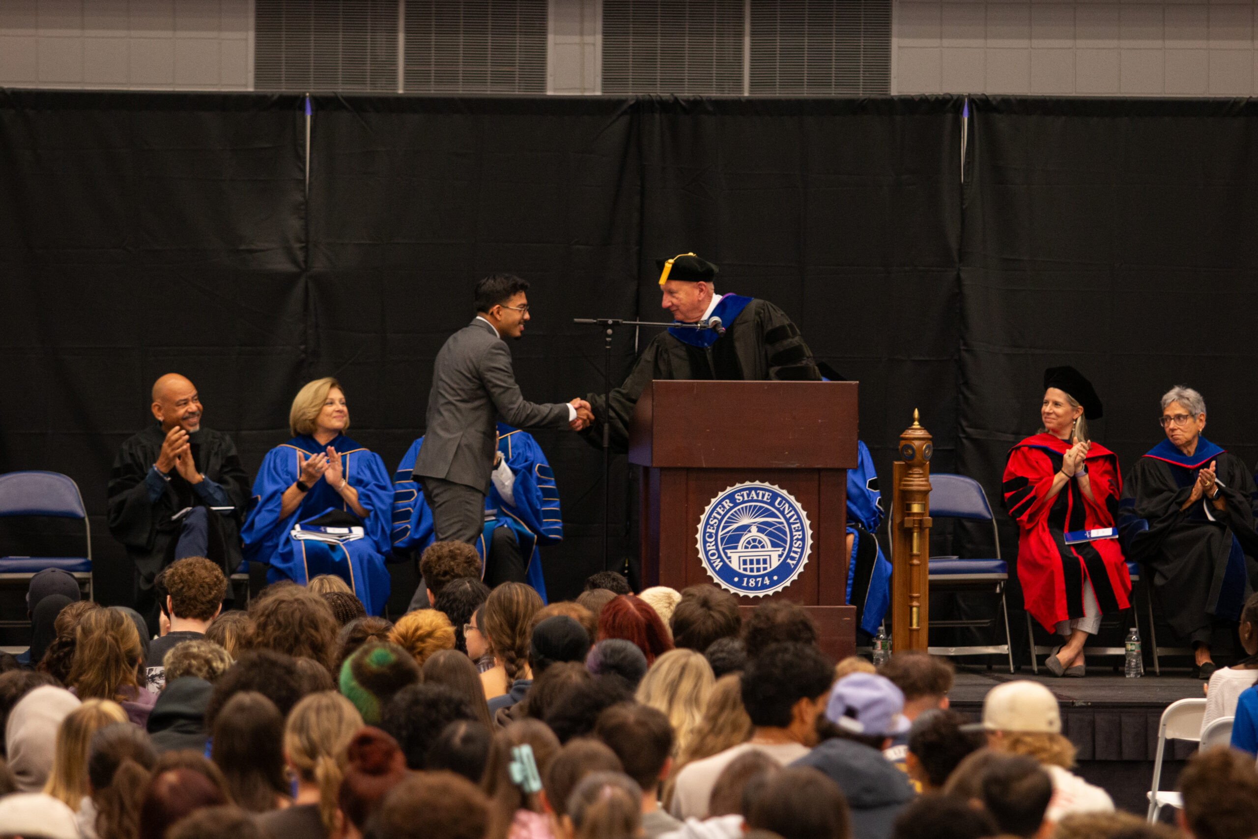 A person in a suit shakes hands with a speaker in academic regalia at a podium during a graduation ceremony, with faculty seated on stage and audience members watching.