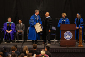A group of faculty in academic regalia on stage at a graduation ceremony; one person is receiving a framed certificate and shaking hands.