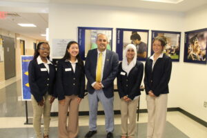 Five people in business attire stand in a hallway, posing for a group photo in front of wall posters and a sign.