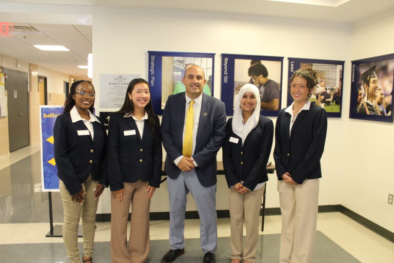Five people in business attire stand in a hallway, posing for a group photo in front of wall posters and a sign.