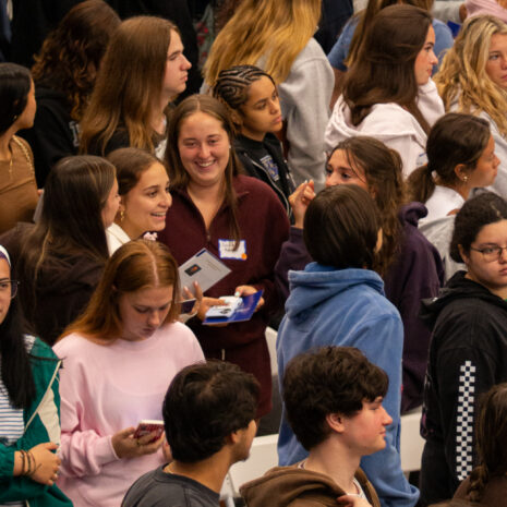 A large group of young people stand closely together indoors, some talking, others looking at phones or holding items.