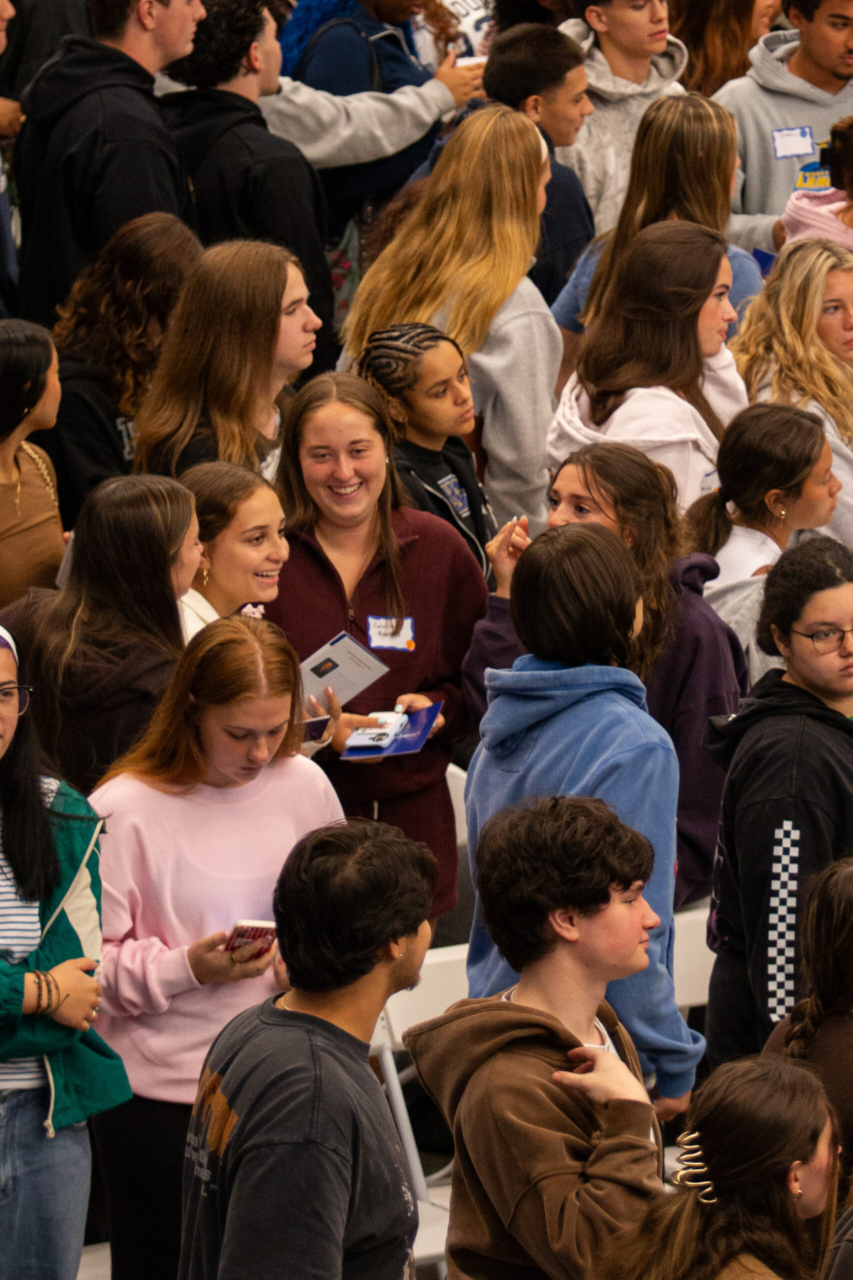 A large group of young people stand closely together indoors, some talking, others looking at phones or holding items.