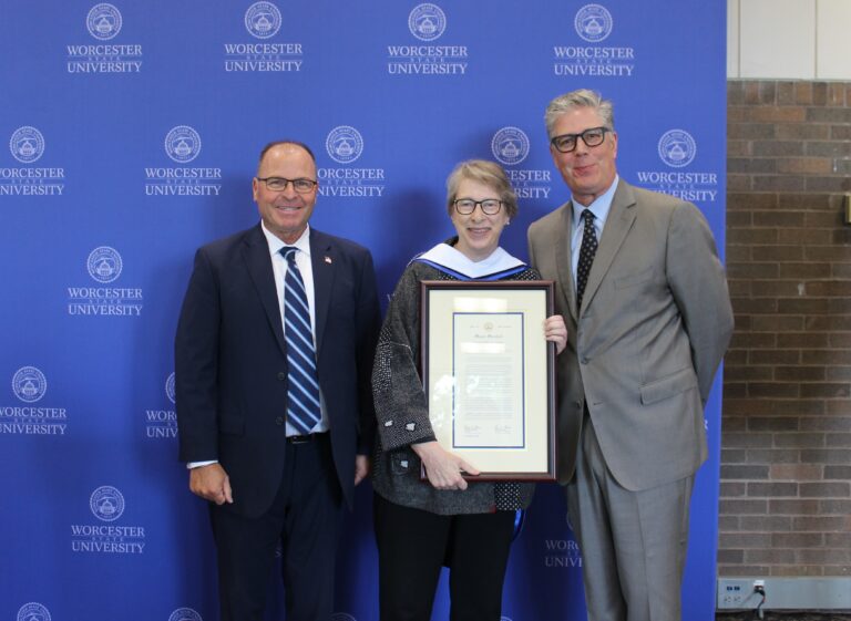 Three people stand in front of a Worcester State University backdrop; the center person holds a framed certificate and wears academic regalia.