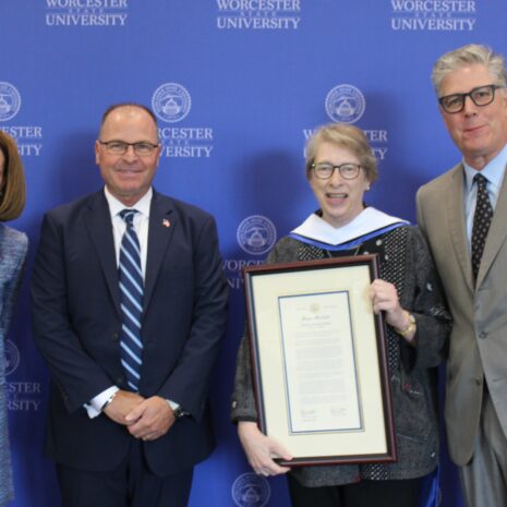 Four people stand in front of a Worcester State University backdrop; one woman holds a framed certificate or award.