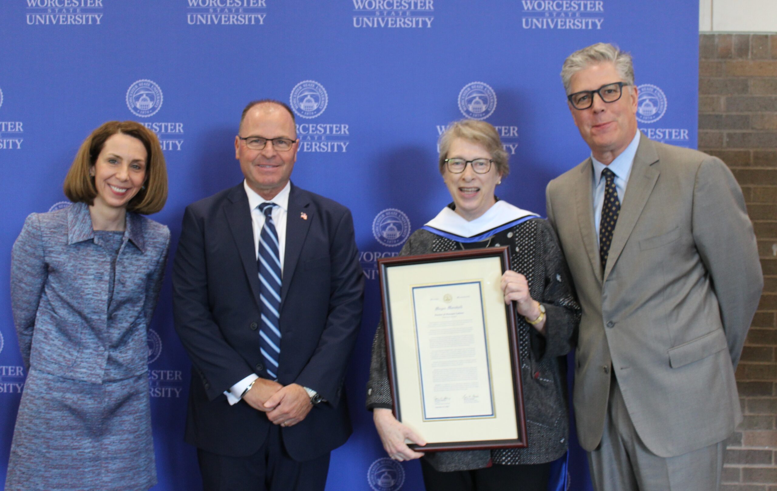 Four people stand in front of a Worcester State University backdrop; one woman holds a framed certificate or award.
