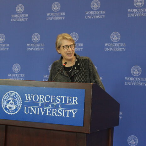 A person stands at a podium with a Worcester State University emblem, speaking in front of a blue backdrop with the university’s name and logo repeatedly printed.