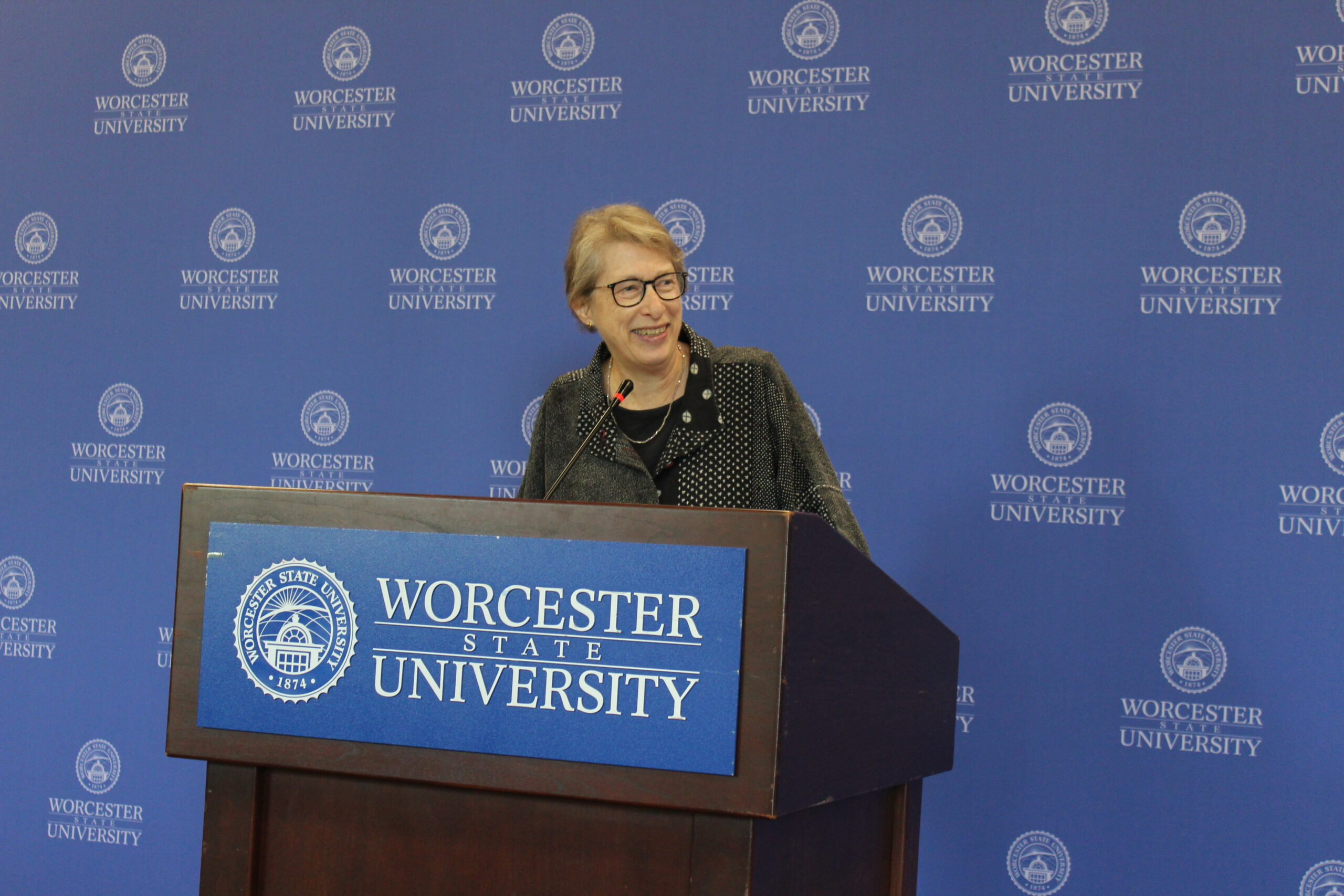 A person stands at a podium with a Worcester State University emblem, speaking in front of a blue backdrop with the university’s name and logo repeatedly printed.