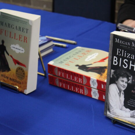 Two books, “Margaret Fuller” and “Elizabeth Bishop: A Miracle for Breakfast,” are displayed on a blue tablecloth, with stacks and partially visible hands nearby.