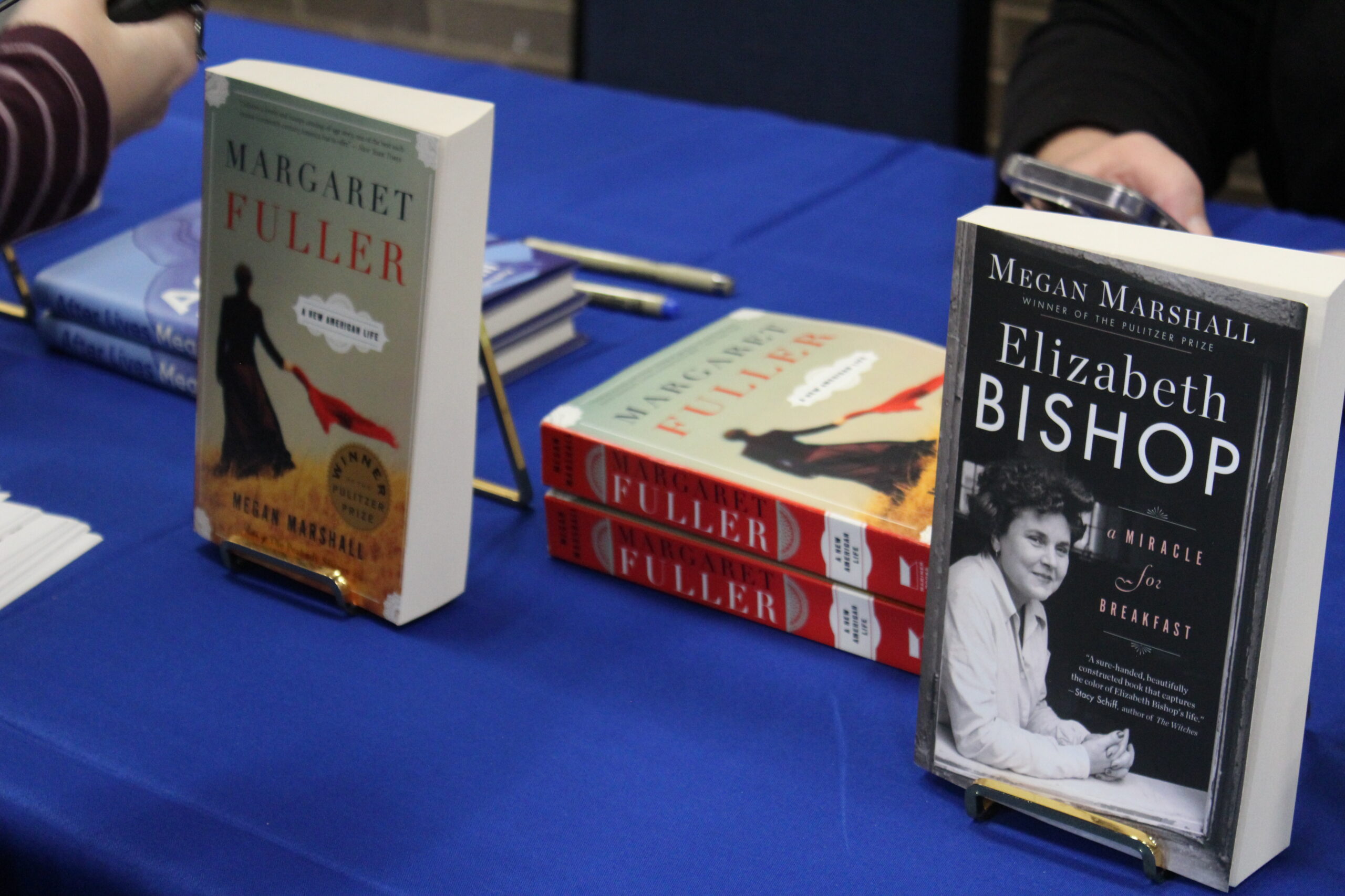 Two books, “Margaret Fuller” and “Elizabeth Bishop: A Miracle for Breakfast,” are displayed on a blue tablecloth, with stacks and partially visible hands nearby.