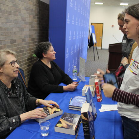 Two authors sit at a table signing books while a woman holding books speaks to them at an indoor event.