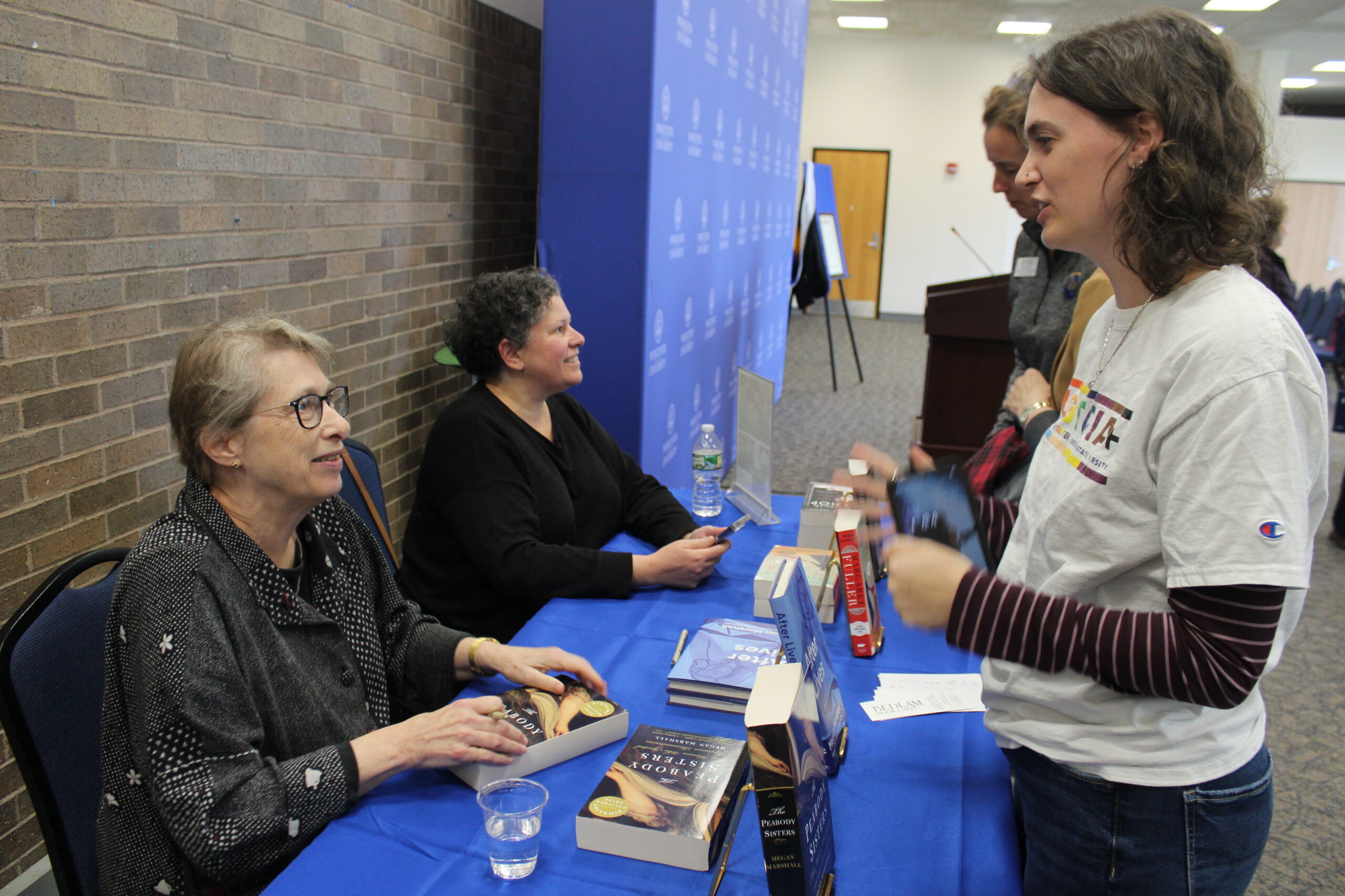 Two authors sit at a table signing books while a woman holding books speaks to them at an indoor event.