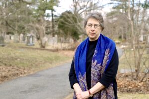 An older woman with short gray hair and glasses stands on a park path, wearing a blue patterned shawl. Trees and gravestones are visible in the background.