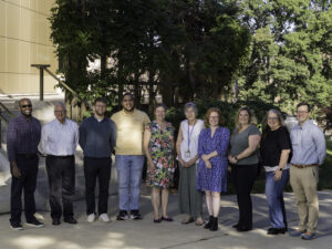 Ten people stand in a row outdoors on a sidewalk, posing for a group photo near trees and a building.