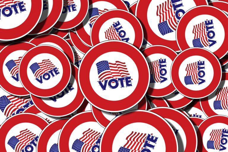 A pile of round buttons with a red border, an American flag, and the word “VOTE” in blue capital letters at the center of each button.