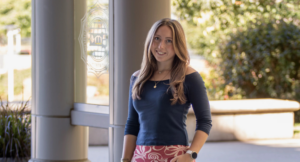 A woman with long brown hair stands outside near a glass door and columns, wearing a navy top and patterned skirt, smiling at the camera.