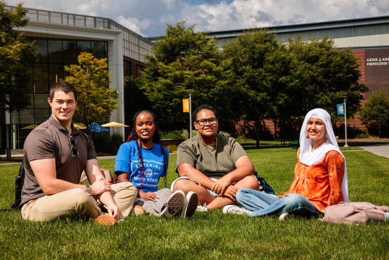 Four students sit together on a grassy campus lawn in front of a university building.