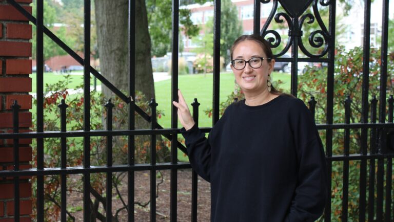 A woman wearing glasses and a black sweatshirt stands with her hand on a black metal gate.