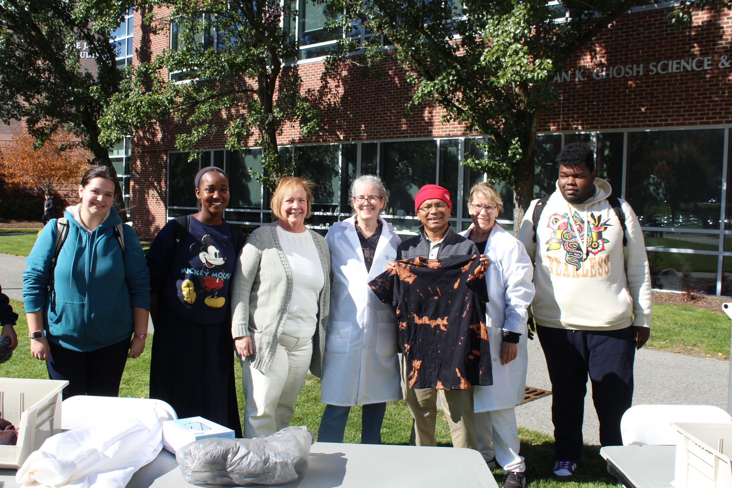 A group of students and faculty members standing in front of a brick science building, with one person holding up a black tie-dye t-shirt.