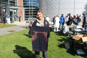 A smiling young woman stands on a lawn holding up a black t-shirt with a brown bleached pattern, with people and building entrances in the background.