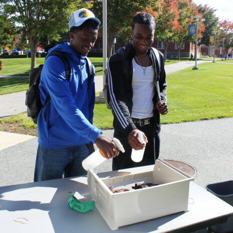 Two young men standing at an outdoor table spray a plastic container filled with items, while another person stands nearby.