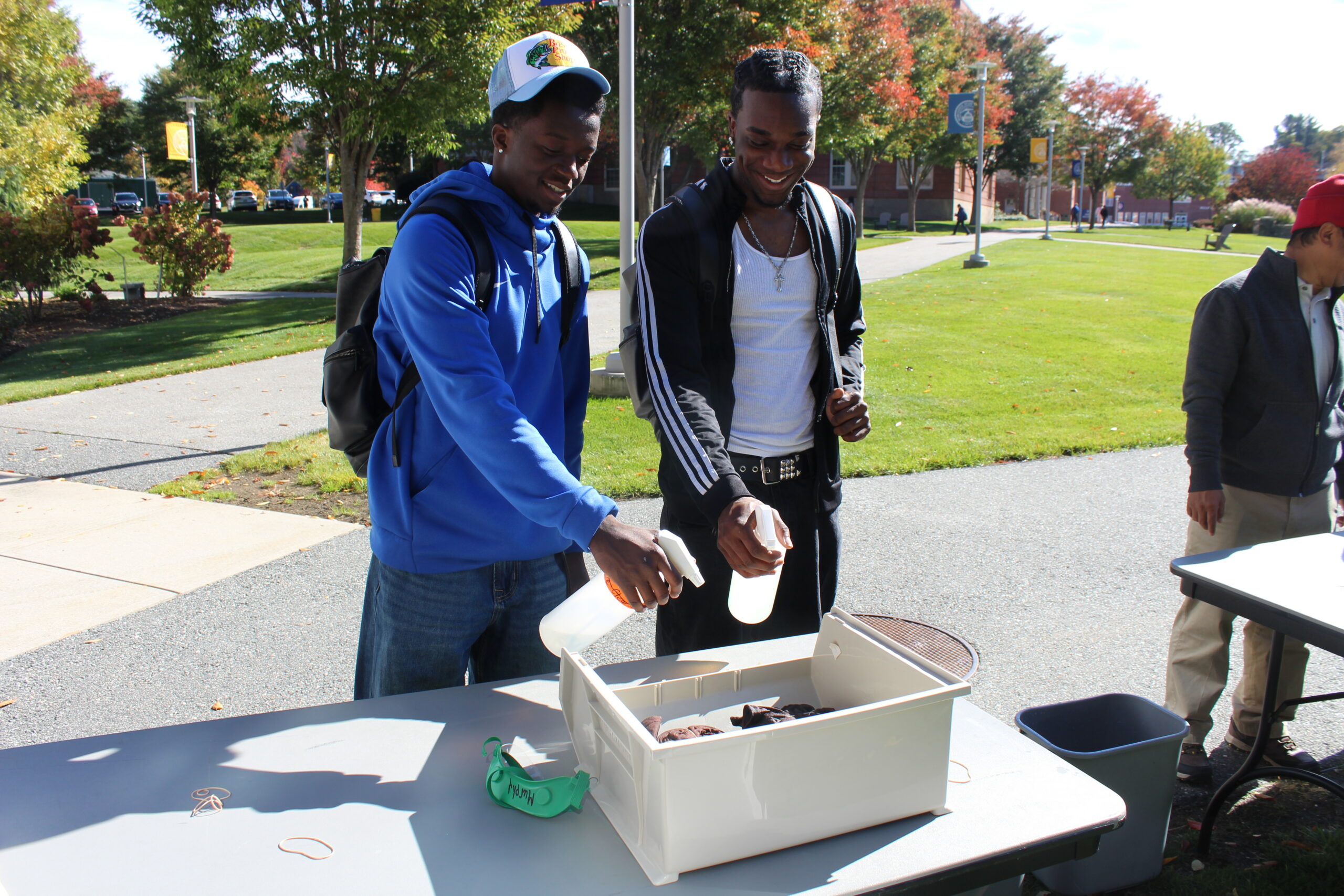 Two young men standing at an outdoor table spray a plastic container filled with items, while another person stands nearby.