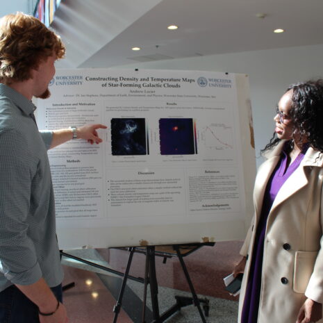 A man pointing to a scientific research poster while speaking with a woman in an indoor hallway.