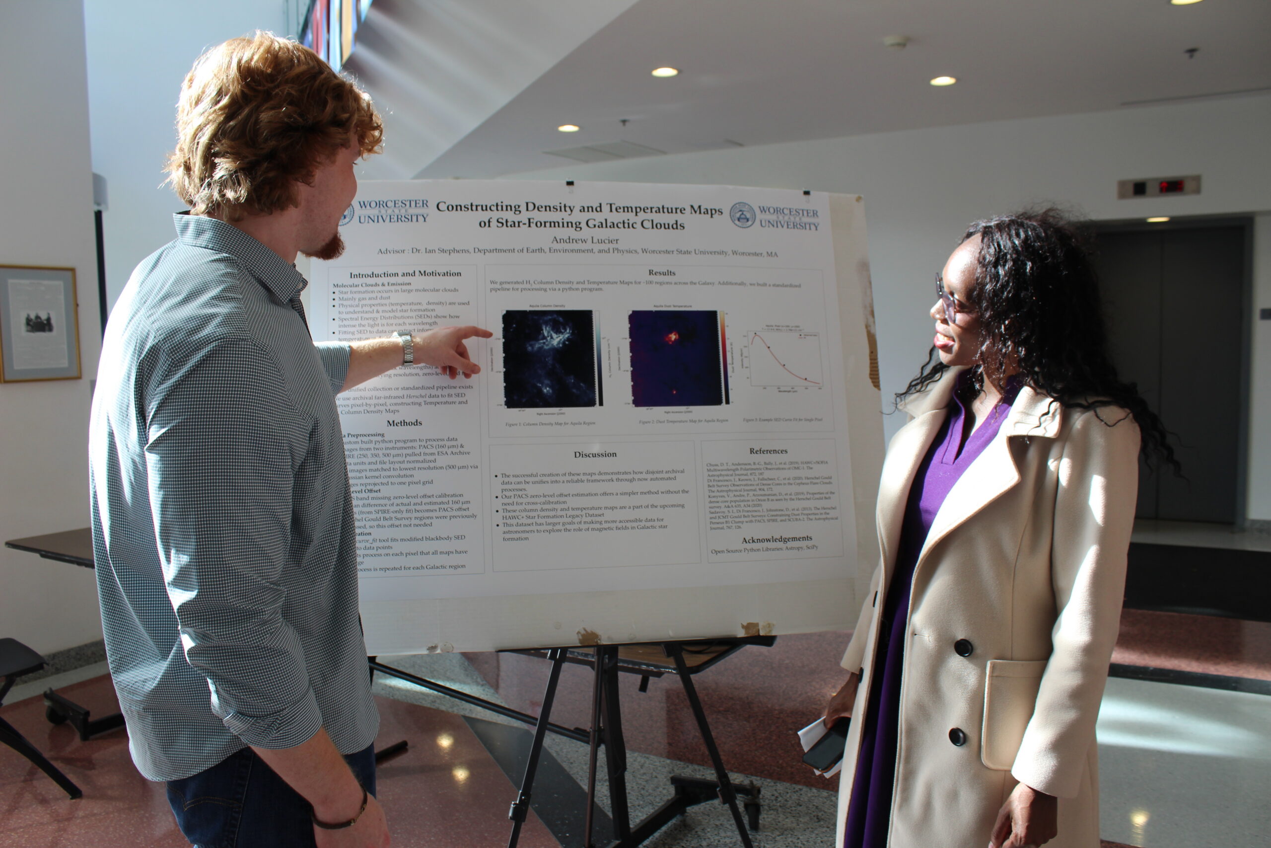A man pointing to a scientific research poster while speaking with a woman in an indoor hallway.