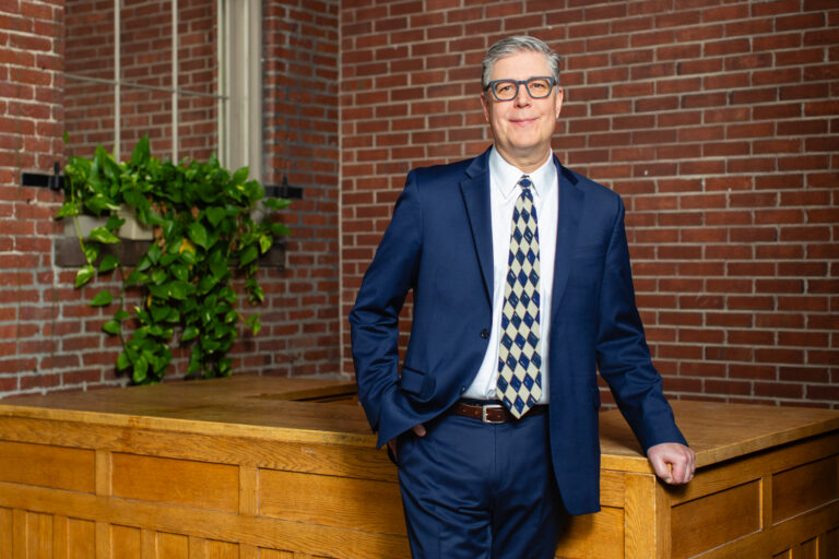 A man in a navy blue suit and patterned tie standing in front of a wooden desk against a brick wall.