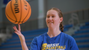 A young woman wearing a WSU Basketball t-shirt spins a basketball on her finger in a gymnasium.