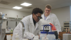 A student looks through a microscope in a laboratory while a professor observes.