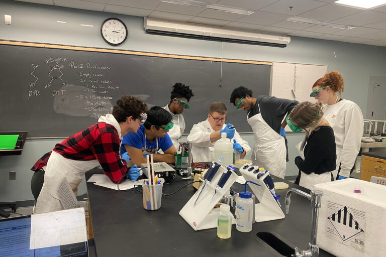 A group of students in lab coats and safety goggles and a professor work together at a lab table with scientific equipment in a classroom.