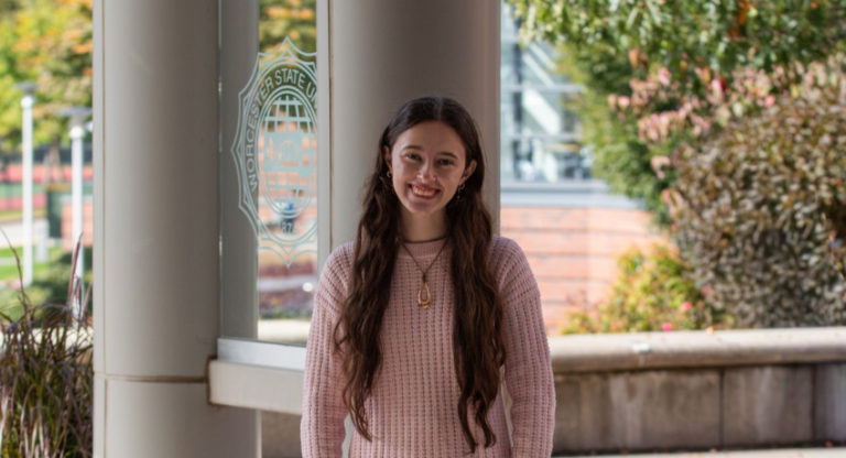 A young woman with long hair smiling in front of a window featuring the Worcester State University logo.